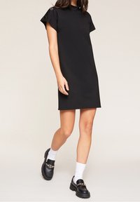 Schwarzes Shiftkleid mit kurzen Ärmeln und Knopfdetails an den Schultern. Glattes, unstrukturiertes Material; knielanges Design. Kombiniert mit klobigen schwarzen Loafers.