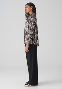 Musterbluse mit schwarzen und beigen Pünktchen, langen Ärmeln und einem Kragen, kombiniert mit schwarzen weiten Hosen und lässigen Turnschuhen.