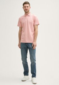 Helles rosa Poloshirt mit einem Kragen und drei Knöpfen, kombiniert mit blauen Jeans und weißen Sneakers. Das Poloshirt hat dezente Akzentverzierungen.