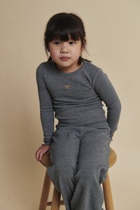 Mladé dievča s tmavými vlasmi, oblečené v sivom dlhorukávovom tričku s výrezom v tvare srdca a sivých nohaviciach, sediace na drevenej stoličke na béžovom pozadí.