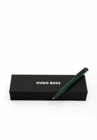 Stylo vert placé à côté d'une boîte noire avec "HUGO BOSS" en lettres blanches ; design épuré avec un clip et une pointe noirs en contraste.
