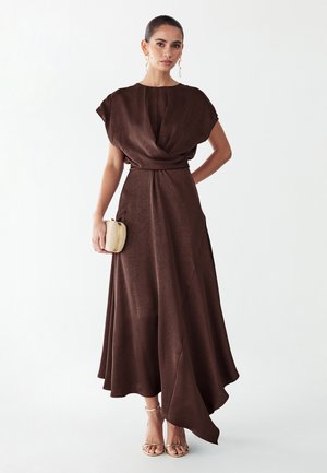 Žena oblečená v tmavohnedej dlhšej šaty bez rukávov s drapovaným živôtikom, drží malú pletenú kabelku a nosí zlaté sandále na remienkoch.