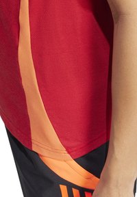 Rotes Sportshirt mit orangefarbenen Akzenten, glatter Textur und entspannter Passform, präsentiert mit schwarzen Shorts, die grafische Designdetails aufweisen.