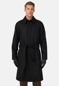 Vīrietis ar garu melnu jostotu mēteli un melnām ādas cimdiem, stāv uz vienkārša gaiša fona.