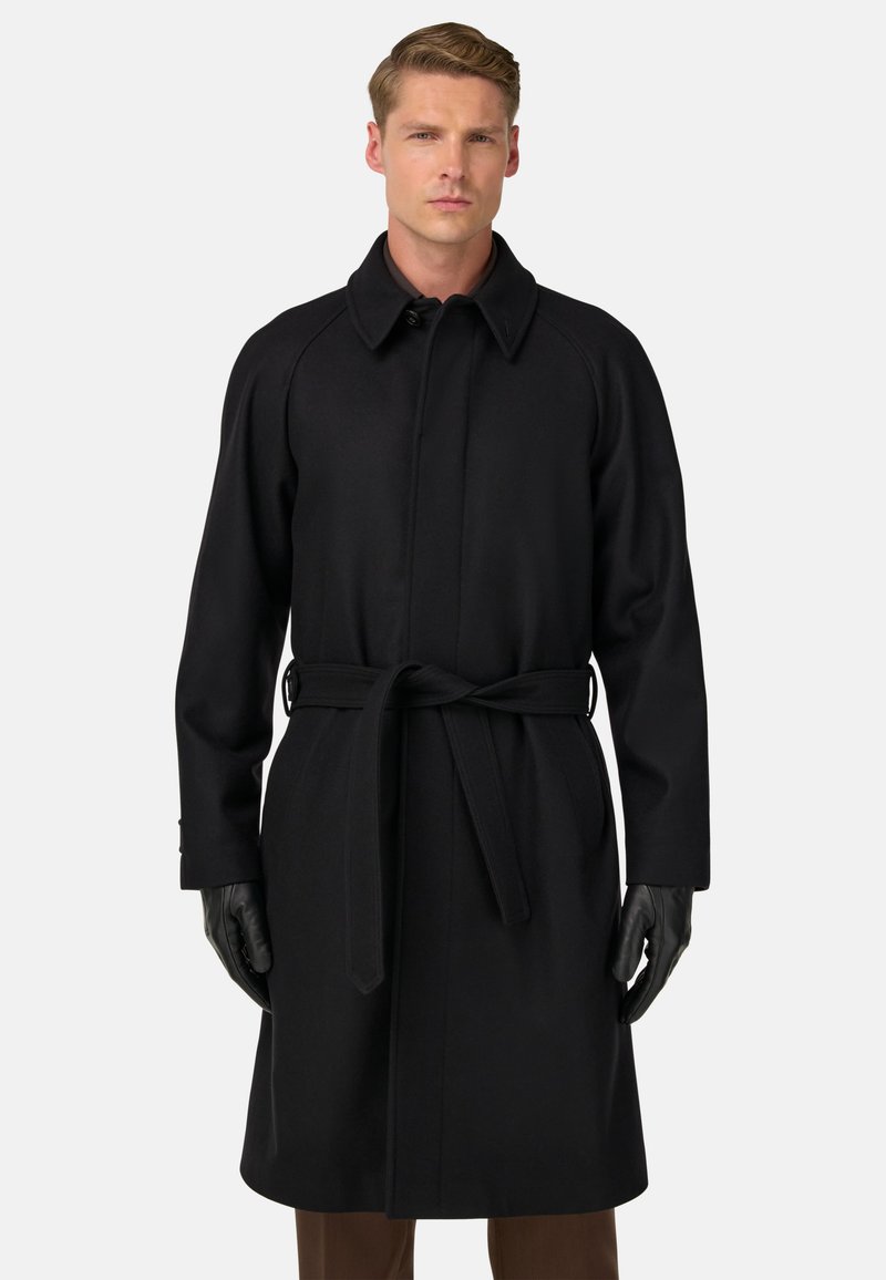 Vīrietis ar garu melnu jostotu mēteli un melnām ādas cimdiem, stāv uz vienkārša gaiša fona.