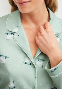 Woman wearing light green pajamas with skiing cat prints, collar, buttons, and white piping along edges, touching her neck.