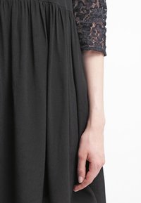 Vestido negro con un cuerpo texturizado y mangas de encaje de tres cuartos; la falda está recogida para una silueta fluida. Mano parcialmente visible.