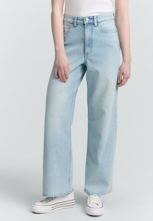 Jeans azzurri a gamba larga in denim, con vita alta e chiusura a bottone, abbinati a sneakers bianche e posizionati su uno sfondo neutro.