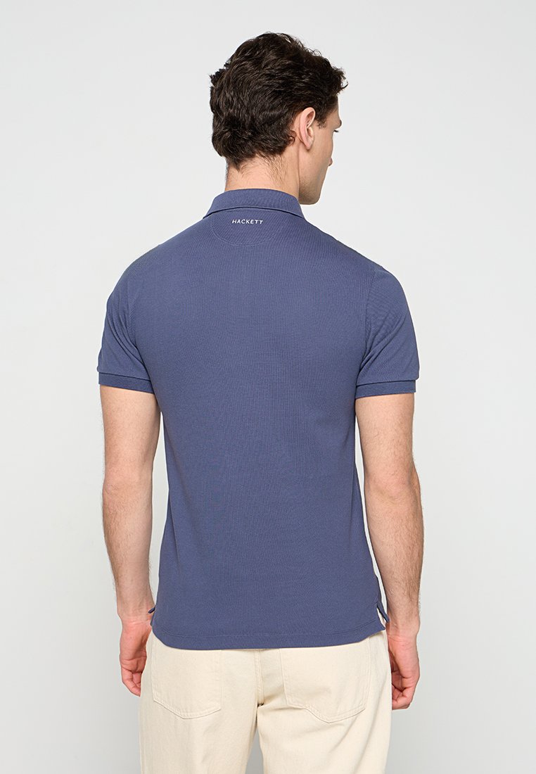 Homme de dos portant un polo bleu ajusté de Hackett et un pantalon beige clair, sur un fond uni clair.