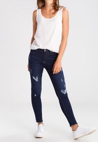 Top sin mangas blanco combinado con jeans ajustados azul oscuro con detalles desgastados y un corte clásico; usado con zapatillas blancas.