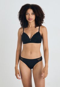 Svart undertøysett med blonder i bølget kant på bralette og matchende truser med blonder. Laget av glatt stoff.