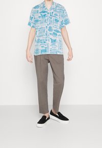 Camicia azzurra a maniche corte con motivi bianchi, abbinata a pantaloni marroni e scarpe slip-on nere con suole bianche, in piedi su uno sfondo uniforme.