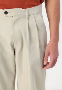 Pantalones plisados en color beige, confeccionados con una tela suave. Presentan un cierre de botón, frente plano y bolsillo lateral, con un ajuste entallado en la cintura.