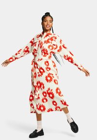 Vestido floral con base blanca, adornado con grandes patrones de flores rojas. Tiene una cintura ceñida con cinturón, mangas largas y un diseño fluido de longitud media.