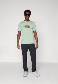 Camiseta verde claro con logo negro y naranja, combinada con pantalones negros. Modelo usando gafas de sol oscuras y zapatillas blancas.