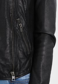 Svart läderjacka med en texturerad yta, med en sneddragkedjeficka och en slät ärmdesign. Metall detaljer inkluderar silverfärgade dragkedjor.