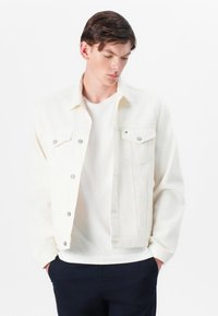 Veste en denim blanche avec fermeture à boutons et poches poitrine. Portée par-dessus un T-shirt blanc, avec un col classique et une coupe décontractée.