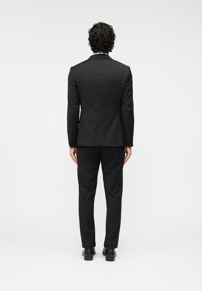 Μαύρο κοστούμι με εφαρμοστή σχεδίαση. Λείο ύφασμα, μονόκουμπο σακάκι, παντελ όνια με στενή γραμμή και γυαλιστερά μαύρα παπούτσια. Μινιμαλιστική αισθητική.