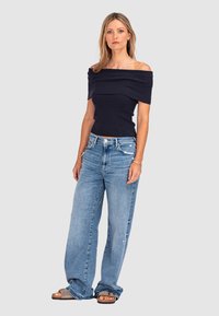 Granatowa bluzka odsłaniająca ramiona z ribbowanym wzorem, jasnoniebieskie dżinsy z szerokimi nogawkami z przetarciami oraz brązowe sandały z szarym detalem w paski.