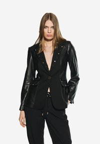 Blazer negro con mangas de cuero, escote en V profundo y tachuelas doradas a lo largo del cuello. Cuenta con un cierre de un solo botón y bolsillos delanteros.