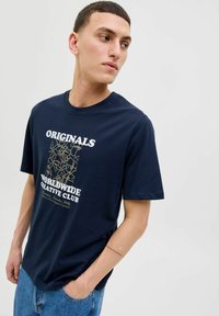 Tamsiai mėlynas medvilninis marškinėliai su trumpomisRankovėmis, kuriuose yra spaudintas dizainas, skelbiantis "ORIGINALS WORLDWIDE CREATIVE CLUB" baltomis ir auksinėmis spalvomis.