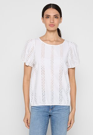 Blouse blanche à manches courtes avec des manches bouffantes, présentant des découpes verticales et un col rond. Associée à un jean bleu.