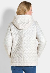 Gepolsterte Steppjacke in hellem Creme mit Kapuze. Verfügt über ein Rautenmuster, seitliche Taschen und gerippte Bündchen. Weicher, glänzender Finish.