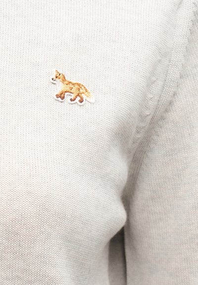 Μικρή κεντημένη πορτοκαλί αλεπού στην περιοχή του στήθους ενός ανοιχτού μπεζ πλεκτού πουλόβερ.