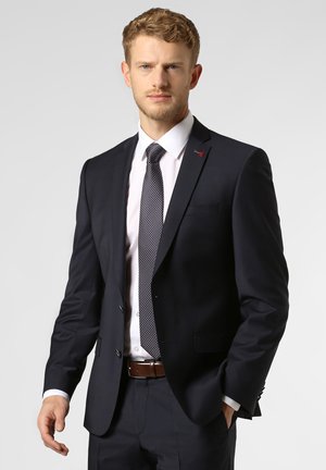 Giacca da abito blu marina con rever a punta, chiusura a bottoni e due tasche anteriori. Abbinata a una camicia bianca e una cravatta a pois. Cintura marrone.