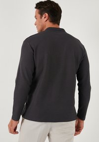 Polo de manga larga, color gris oscuro, con una tela texturizada, corte recto y aberturas laterales, que presenta un cuello sutil y costuras mínimas.