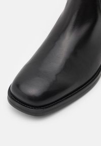 Botte en cuir noir avec une finition lisse et brillante, bout arrondi et détails de coutures discrètes le long de la semelle. Semelle extérieure en caoutchouc plate visible.