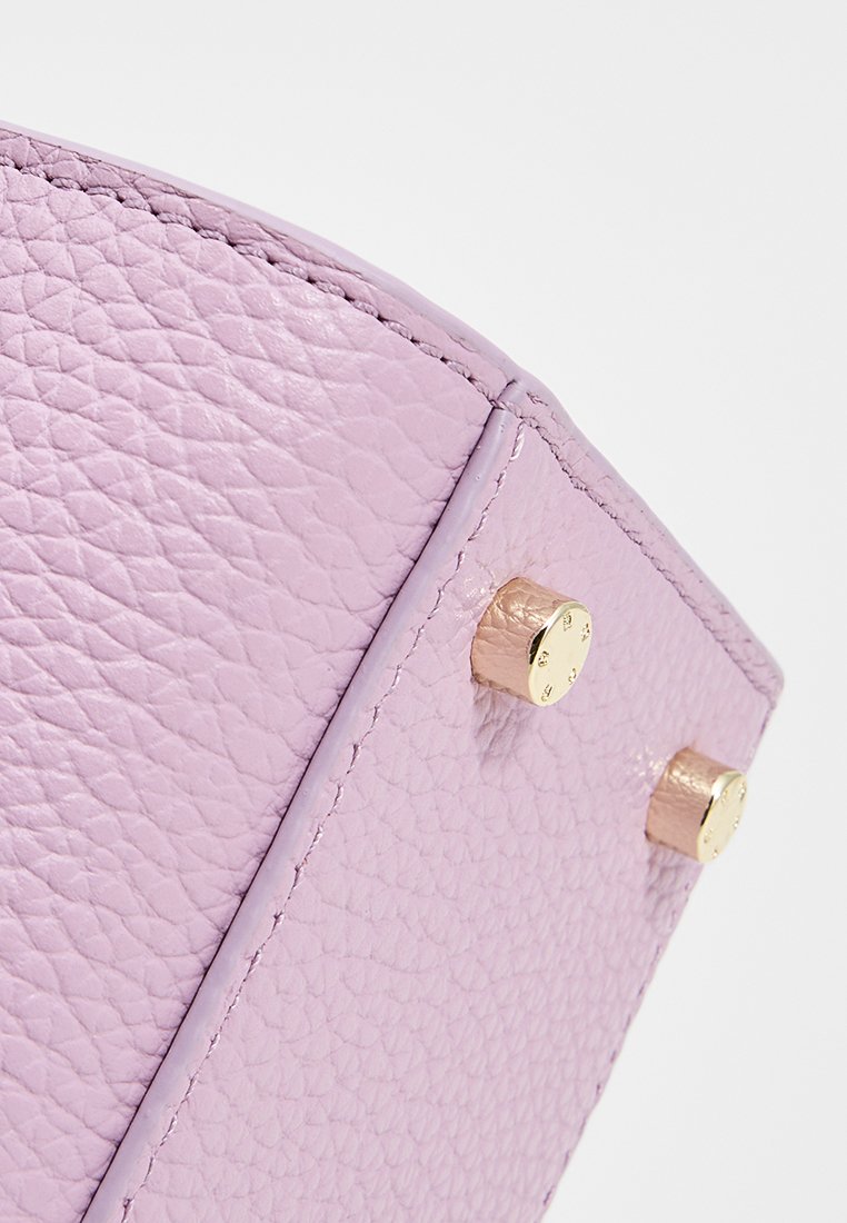 Nahaufnahme einer Ecke einer lila strukturierten Lederhandtasche mit zwei kleinen goldenen Metallnieten und detaillierter Naht auf weißem Hintergrund.