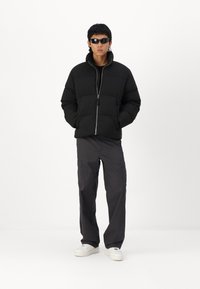 Must must jacket kõrge kaelusega ja tõmblukuga, beež tumedate cargo-pükste ja valgete tossudega. Mudel kannab musti prille.