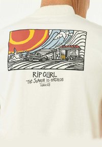 Beige Baumwoll-T-Shirt mit einem Druckdesign eines Autos, einer Tankstelle und einem Sonnenuntergang in kräftigen Rot-, Gelb- und Blautönen. Der Text lautet "RIP CURL DIE SUCHE IST ENDLOS SEIT 69."