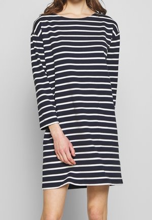 Mujer vestida con un vestido de manga larga a rayas horizontales azul marino y blanco, con escote barco, de pie contra un fondo liso.