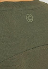 Dunkelgrünes T-Shirt mit rundem Ausschnitt. Glatte Textur und subtile Stichdetails. Verfügt über ein auffälliges kreisförmiges Logo im oberen Rückenbereich.