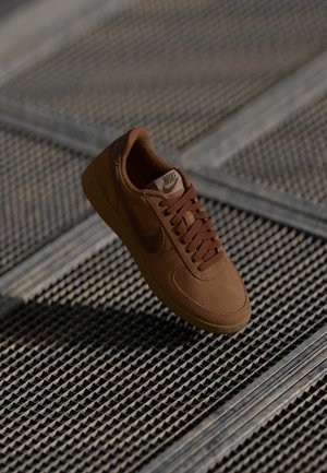 Brązowy but sportowy Nike z sznurowadłami unoszący się ponad teksturowanym metalowym rusztem na tle miękkiego oświetlenia.