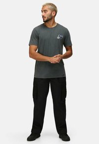 Graues Baumwoll-T-Shirt mit kurzen Ärmeln und rundem Hals, das mit einem kleinen blauen Grafik-Logo versehen ist. Kombiniert mit schwarzen Cargo-Hosen.