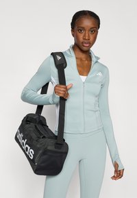 Ανοιχτό μπλε αθλητικό μπουφάν με φερμουάρ, φορεμένο με αντίστοιχο κολάν· μαύρη τσάντα duffel με λευκό λογότυπο Adidas και ανθεκτικό ιμάντα ώμου.