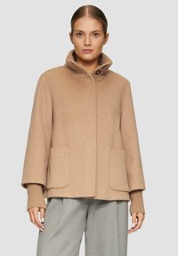 Beige Wollmischungs-Mantel mit hohem Kragen, ausgestattet mit zwei vorderen Taschen und gerippten Strickbündchen, präsentiert in einer lässigen Silhouette.