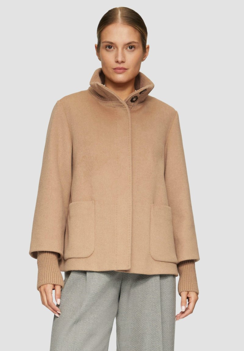 Beige Wollmischungs-Mantel mit hohem Kragen, ausgestattet mit zwei vorderen Taschen und gerippten Strickbündchen, präsentiert in einer lässigen Silhouette.