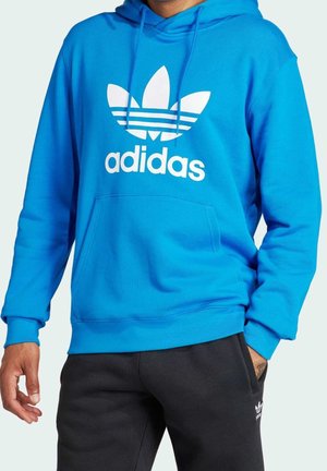 Mężczyzna noszący jasnoniebieską bluzę z kapturem Adidas z białym logo oraz czarne spodnie dresowe Adidas, z ręką w kieszeni na jasnym tle.