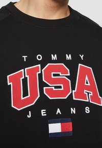 Gros plan d’un sweat-shirt noir avec le texte « TOMMY USA JEANS » et un petit logo rectangulaire rouge, blanc et bleu brodé sous les lettres.