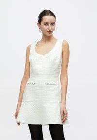 Biała sukienka bez rękawów o fakturowanej powierzchni, z dekoltem w kształcie litery U, rozkloszowanym dołem i zdobionymi kieszeniami. Modelka ma na sobie czarne rajstopy.