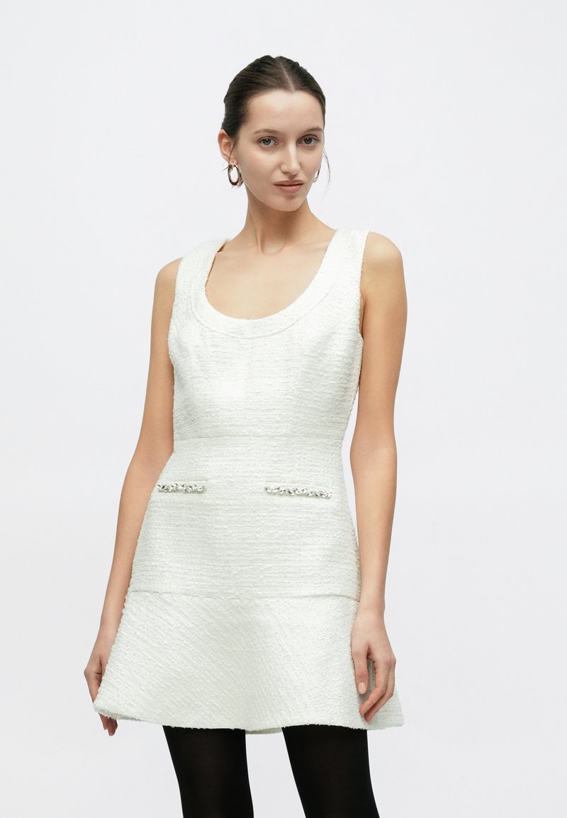 Biała sukienka bez rękawów o fakturowanej powierzchni, z dekoltem w kształcie litery U, rozkloszowanym dołem i zdobionymi kieszeniami. Modelka ma na sobie czarne rajstopy.