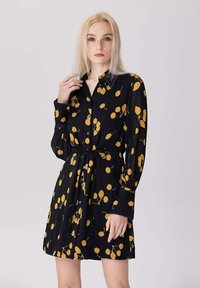 Femme blonde portant une robe noire à boutons ornée de motifs de cerises jaunes et avec une ceinture à la taille, se tenant devant un fond neutre.