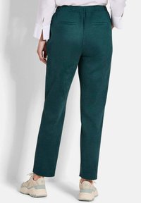 Elastische taillebroek van diep teal stof met een gladde textuur, voorzien van twee achterzakken en een rechte pasvorm. Gemaakt met beige sneakers.