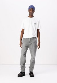 Λευκό κοντομάνικο t-shirt με μαύρο λογότυπο, ανοιχτό γκρι τζιν και μαύρες κορδονάτες μπότες. Το μοντέλο φοράει ένα μπλε ριμπ καπέλο. Απλό φόντο.