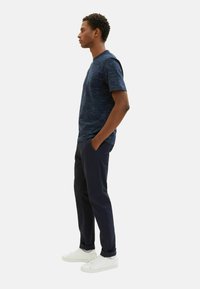 Mörkblå strukturerad t-shirt med kort ärm, kombinerad med mörka byxor och vita sneakers, sedd från sidan.