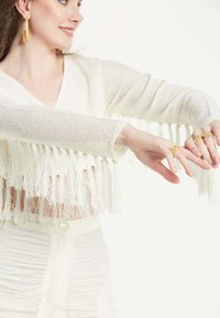 Weißes gestricktes Cropped-Top mit Fransenverzierung am Saum, kombiniert mit einem hellen, strukturierten Rock. Modell trägt goldene Ringe und Ohrringe.